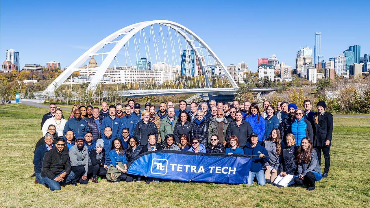 Un grand groupe d'employés de Tetra Tech tenant une bannière Tetra Tech dans un parc avec un horizon urbain et un pont en arrière-plan