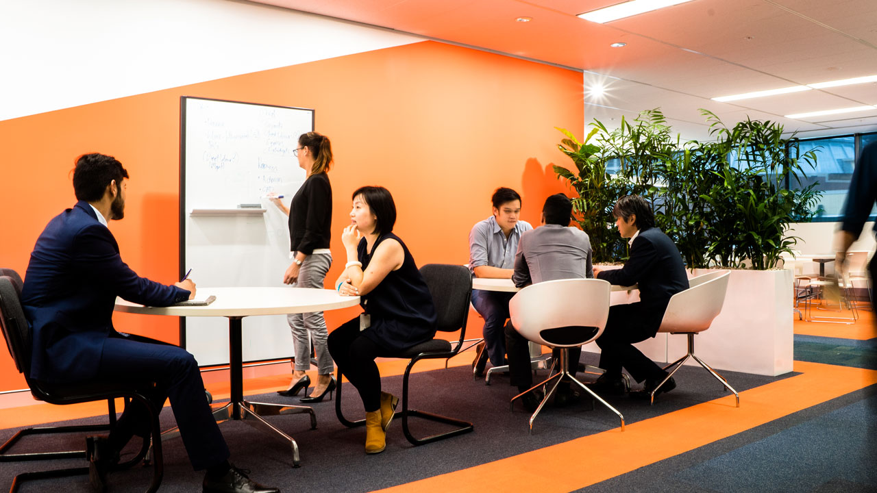 Une femme prend des notes sur un tableau blanc, et d’autres personnes sont assises en groupe autour de tables dans un espace de travail collaboratif informel dont les murs sont de couleur orange