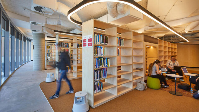 Dans une bibliothèque universitaire, un étudiant marche entre des rayons, au deuxième plan. Au premier plan, des étudiants sont assis et utilisent des ordinateurs portatifs