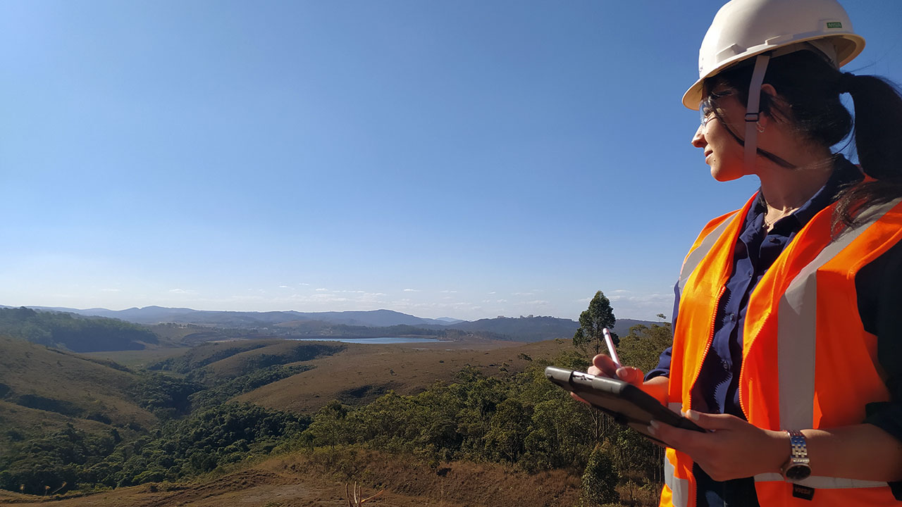 Une employée de Tetra Tech équipée d’un gilet et d’un casque de sécurité tient une planchette à pince et observe le relief depuis le sommet d’une colline
