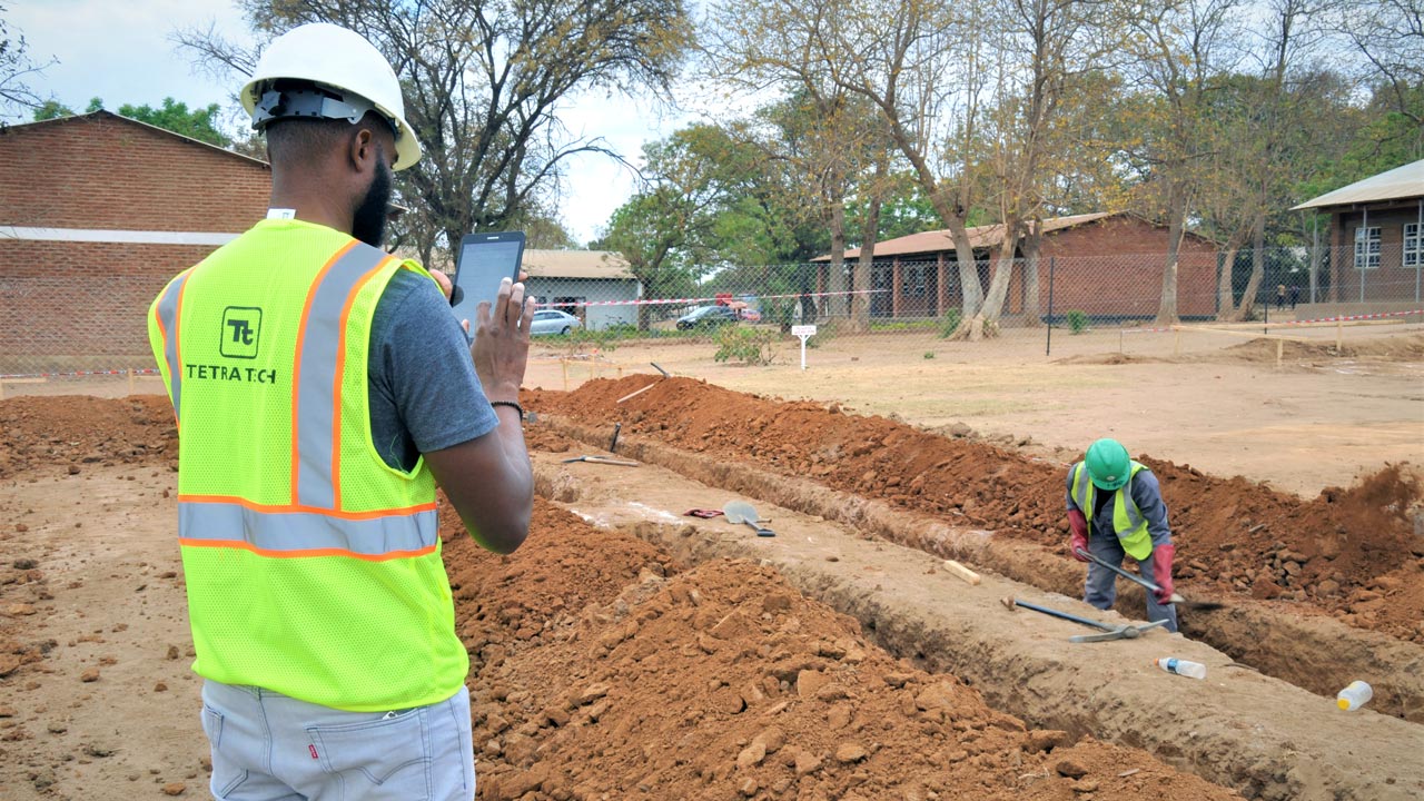 Un technicien de Tetra Tech portant un gilet à haute visibilité tient une tablette et observe un ouvrier en train de creuser une tranchée au Malawi