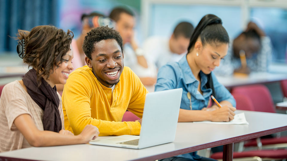 Trois étudiants noirs sont assis à une table; deux d’entre eux travaillent sur un ordinateur portatif, tandis que le troisième est en train de prendre des notes sur un papier