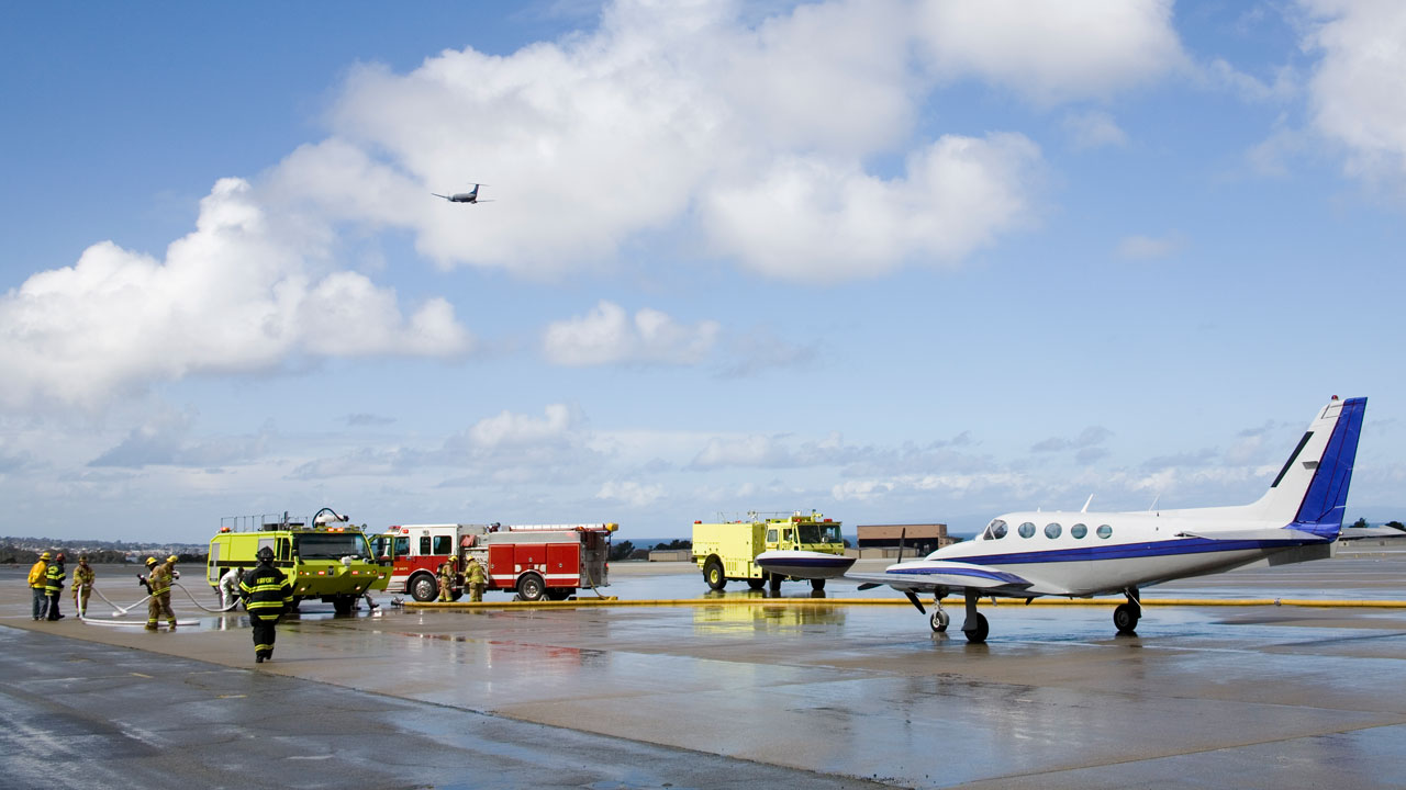 Camions de pompiers et pompiers sur l’aire de trafic avec un petit avion sous un grand ciel bleu avec des nuages floconneux