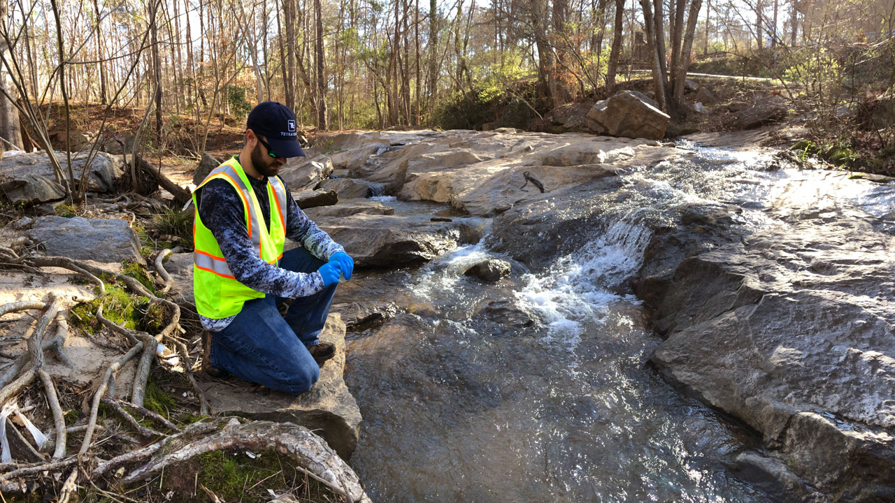 Un technicien portant un équipement de sécurité est agenouillé sur un rocher et se prépare à prélever des échantillons d’eau de rivière