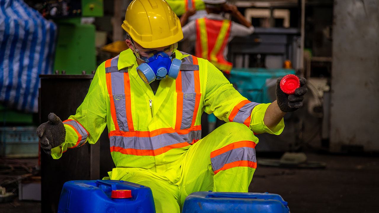 Un ingénieur portant une combinaison de sécurité, des gants noirs, un masque à gaz et un casque de sécurité contrôle un bidon de produits chimiques dans une usine