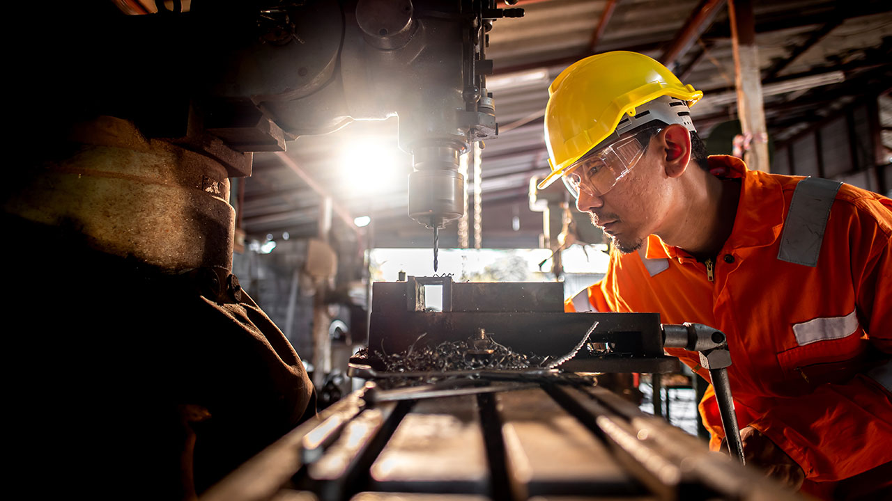 Un technicien portant des vêtements de travail de couleur orange et des lunettes de sécurité se tient à côté d’une perceuse et utilise des forets métalliques