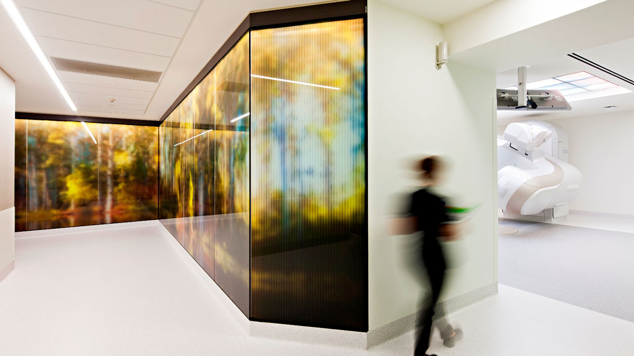 Un professionnel de la santé marche dans un couloir constitué d’une paroi de verre colorée en direction d’une salle ouverte de tomographie par ordinateur