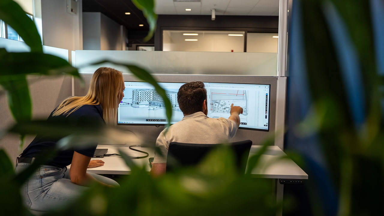 Une collègue et un collègue sont assis un à côté de l'autre à un bureau et ils regardent un écran d'ordinateur. Le collègue pointe un plan affiché à l'écran